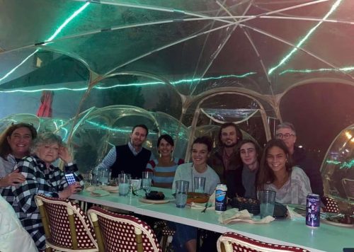 A group of people dining together under a transparent tent, enjoying food and drinks in a cozy setting.