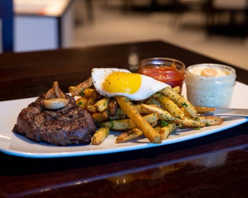 A plate featuring a grilled steak, fried egg, seasoned fries, and dipping sauces.