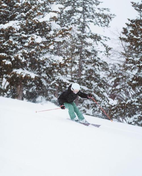 A skier in green pants and a black jacket navigates a snowy slope amidst tall trees. Snow is falling around them.