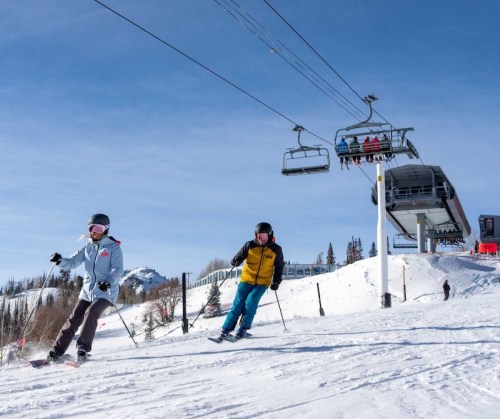 Two skiers navigate a snowy slope under a clear blue sky, with a ski lift in the background.