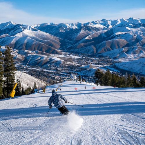 A skier descends a snowy slope with mountains in the background under a clear blue sky.