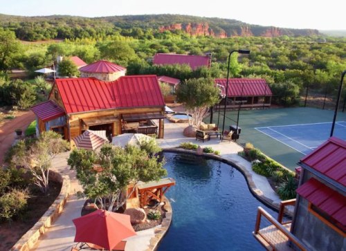 Aerial view of a rustic property with a pool, tennis court, and red-roofed cabins surrounded by greenery.