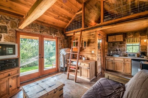 Cozy wooden cabin interior with a loft, large windows, and rustic furniture, overlooking a green outdoor area.