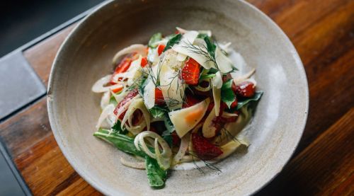 A fresh salad with mixed greens, sliced onions, red peppers, and topped with shaved cheese and herbs in a bowl.