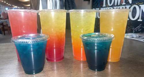 A variety of colorful drinks in clear cups, including red, yellow, orange, and blue, arranged on a wooden table.