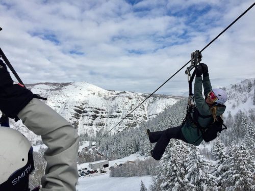 A person zip-lining over a snowy landscape with mountains and trees in the background.