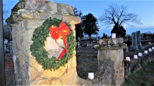 A stone monument adorned with a wreath and bow, set in a cemetery with glowing lanterns lining the path at dusk.