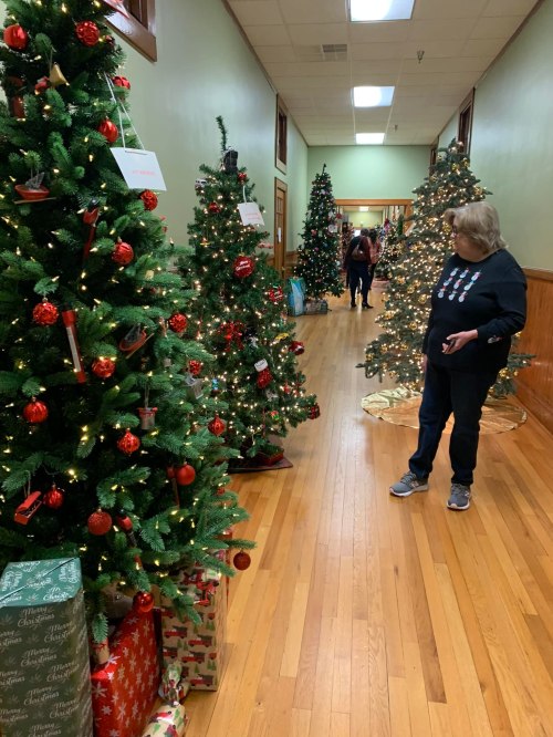A hallway lined with decorated Christmas trees and a woman admiring one, with people in the background.