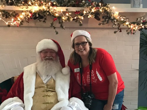 A smiling woman in a red shirt poses with Santa Claus in front of a festive fireplace decorated with lights and greenery.