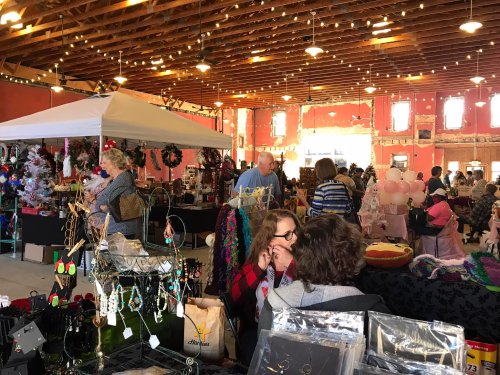 A bustling indoor market with vendors, shoppers, and festive decorations, showcasing handmade crafts and holiday items.