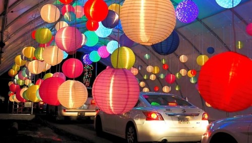 Colorful lanterns hang from a canopy, illuminating a pathway where cars are parked below.