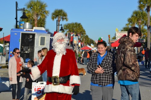 A festive street scene with Santa Claus, families, and vendors, palm trees in the background under a clear blue sky.