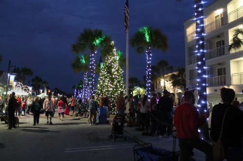 A festive street scene at night, with palm trees lit up and a crowd enjoying holiday decorations and activities.