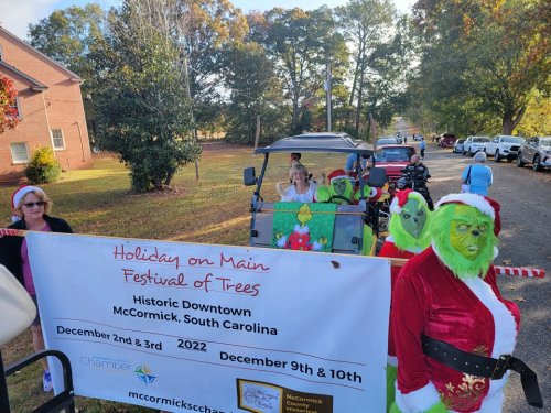 A festive parade scene with people in Grinch costumes and a banner for the Holiday on Main Festival of Trees in McCormick, SC.