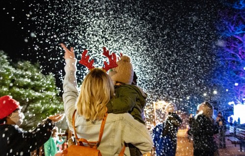 A woman holds a child with reindeer antlers as they enjoy falling snow at a festive outdoor event.