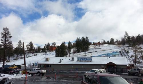 A snowy landscape with a ski resort building and ski slopes in the background under a partly cloudy sky.