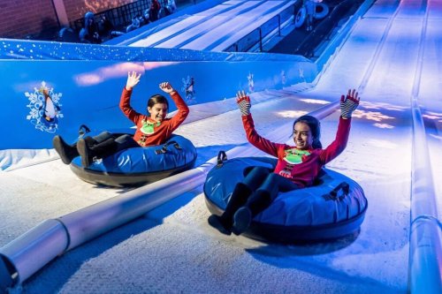 Two children in festive sweaters joyfully sledding down a snowy slope, waving their hands in excitement.