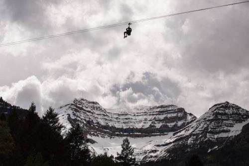 A person zip-lining against a backdrop of snow-capped mountains and cloudy skies.