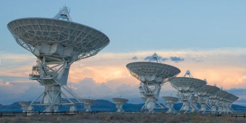 A row of large white satellite dishes against a cloudy sky, set in a desert landscape.