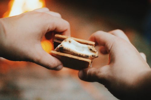 A person holding a s'more with toasted marshmallow and chocolate, with a campfire glowing in the background.