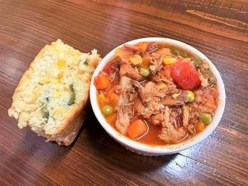 A bowl of hearty stew with meat and vegetables, served alongside a slice of cornbread on a wooden table.