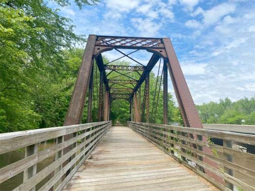 A metal truss bridge spans a tree-lined path under a blue sky with scattered clouds.