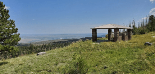 A scenic view of a grassy area with a stone shelter overlooking a valley and distant mountains under a clear blue sky.