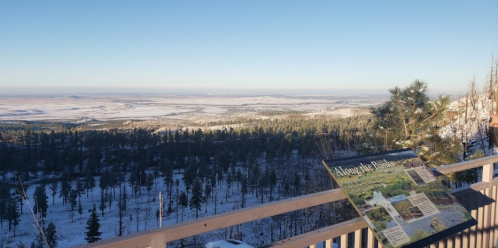 A snowy landscape with distant hills and trees, featuring an informational sign in the foreground.