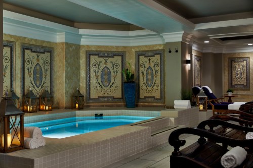 A serene spa interior featuring a mosaic wall, a small pool, and wooden lounge chairs with rolled towels.
