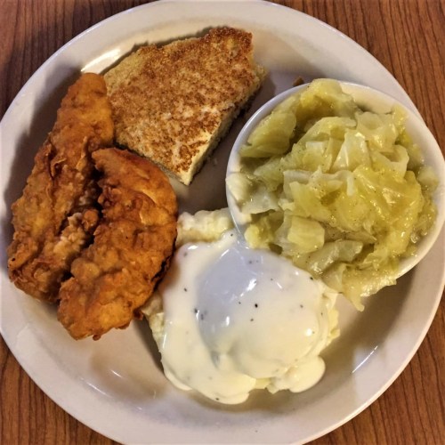 A plate of fried chicken, mashed potatoes with gravy, sautéed cabbage, and a piece of cornbread.