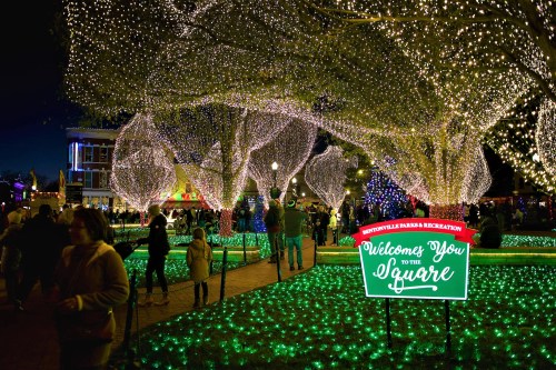 A festive square illuminated with colorful lights, featuring a welcome sign and people enjoying the holiday atmosphere.