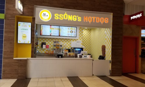 A hotdog stand with a bright sign, menu displays, and a counter in a food court setting.