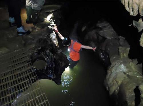 A person in an orange shirt wades through water in a cave, with others observing nearby. Headlamps illuminate the scene.
