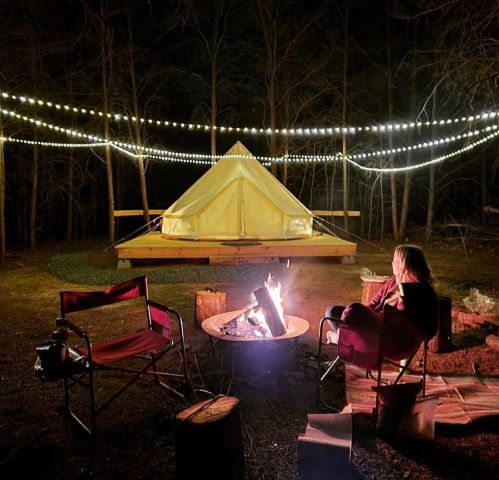 A cozy campsite at night with a glowing tent, string lights, a campfire, and a person sitting nearby.