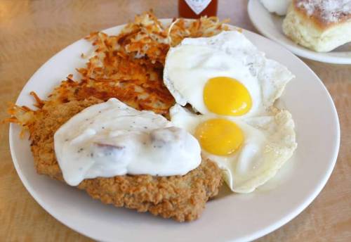 A plate of crispy fried chicken topped with white gravy, served with two sunny-side-up eggs and hash browns.