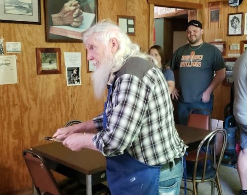 An elderly man with a long white beard stands in a rustic room, holding tools, while others watch in the background.