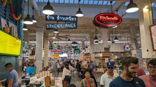 A bustling market scene with various food stalls and colorful signs, filled with people enjoying the atmosphere.