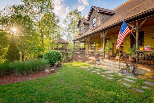 A charming yellow house with a porch, surrounded by greenery and an American flag, under a sunny sky.