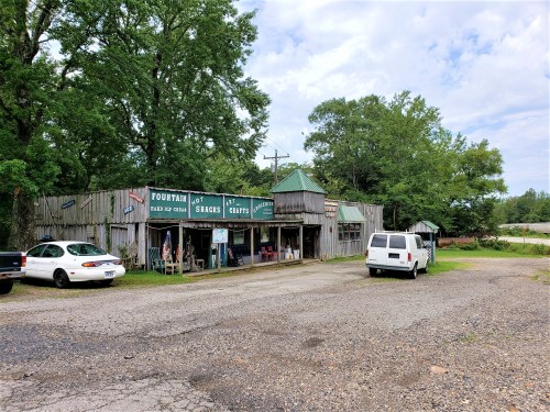 A rustic roadside shop with a sign for ice cream and snacks, surrounded by trees and parked cars.