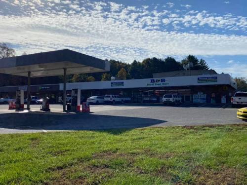 A convenience store with a gas station, surrounded by vehicles and green grass under a cloudy blue sky.