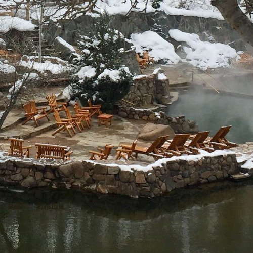 A snowy landscape featuring wooden chairs arranged around a stone patio near a steaming body of water.