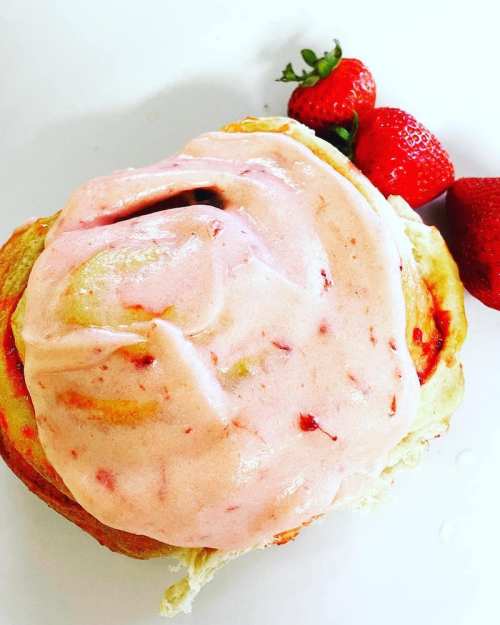 A cinnamon roll topped with pink strawberry frosting, accompanied by fresh strawberries on a white plate.
