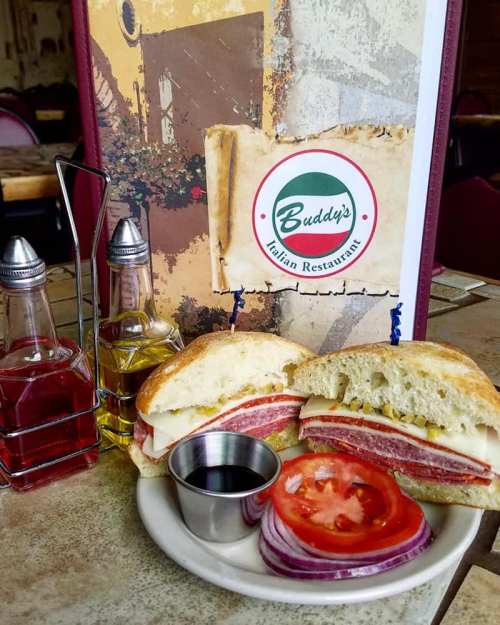 A sandwich with layers of meats and cheese, served with tomato and onion slices, alongside oil and vinegar bottles.