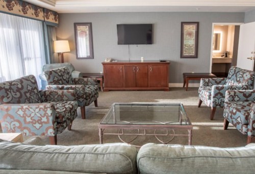 A spacious hotel living area with patterned chairs, a glass coffee table, and a TV on the wall.