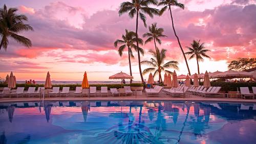 A serene poolside scene at sunset, with palm trees, lounge chairs, and a colorful sky reflecting in the water.