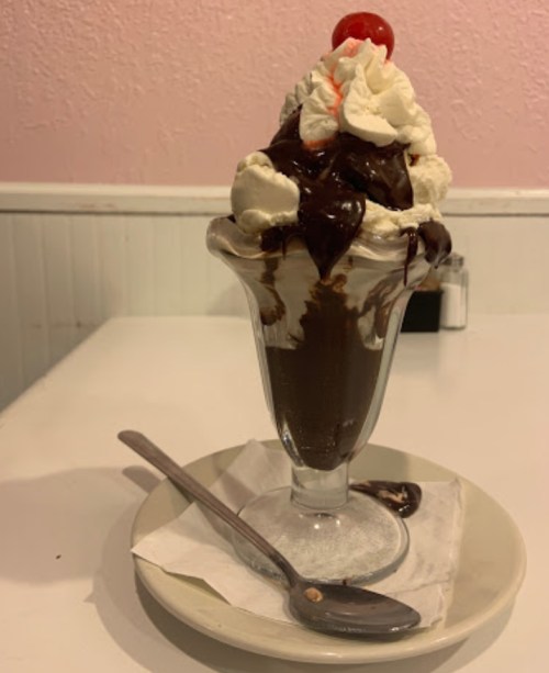 A tall glass sundae with chocolate sauce, whipped cream, and a cherry on top, served on a plate with a spoon.