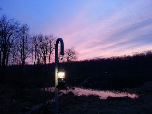A lantern hangs by a calm pond at sunset, with trees silhouetted against a colorful sky.