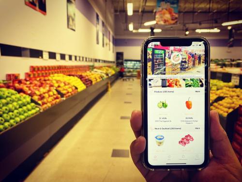 A hand holds a smartphone displaying a grocery app in a colorful produce aisle filled with fruits and vegetables.