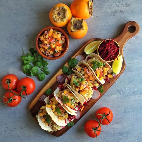 A wooden platter with tacos topped with mango salsa, cilantro, and radishes, alongside tomatoes and persimmons.