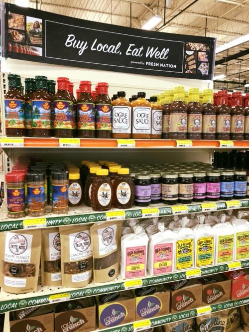 A grocery shelf displaying various local sauces, spices, and condiments with a sign that reads "Buy Local. Eat Well."